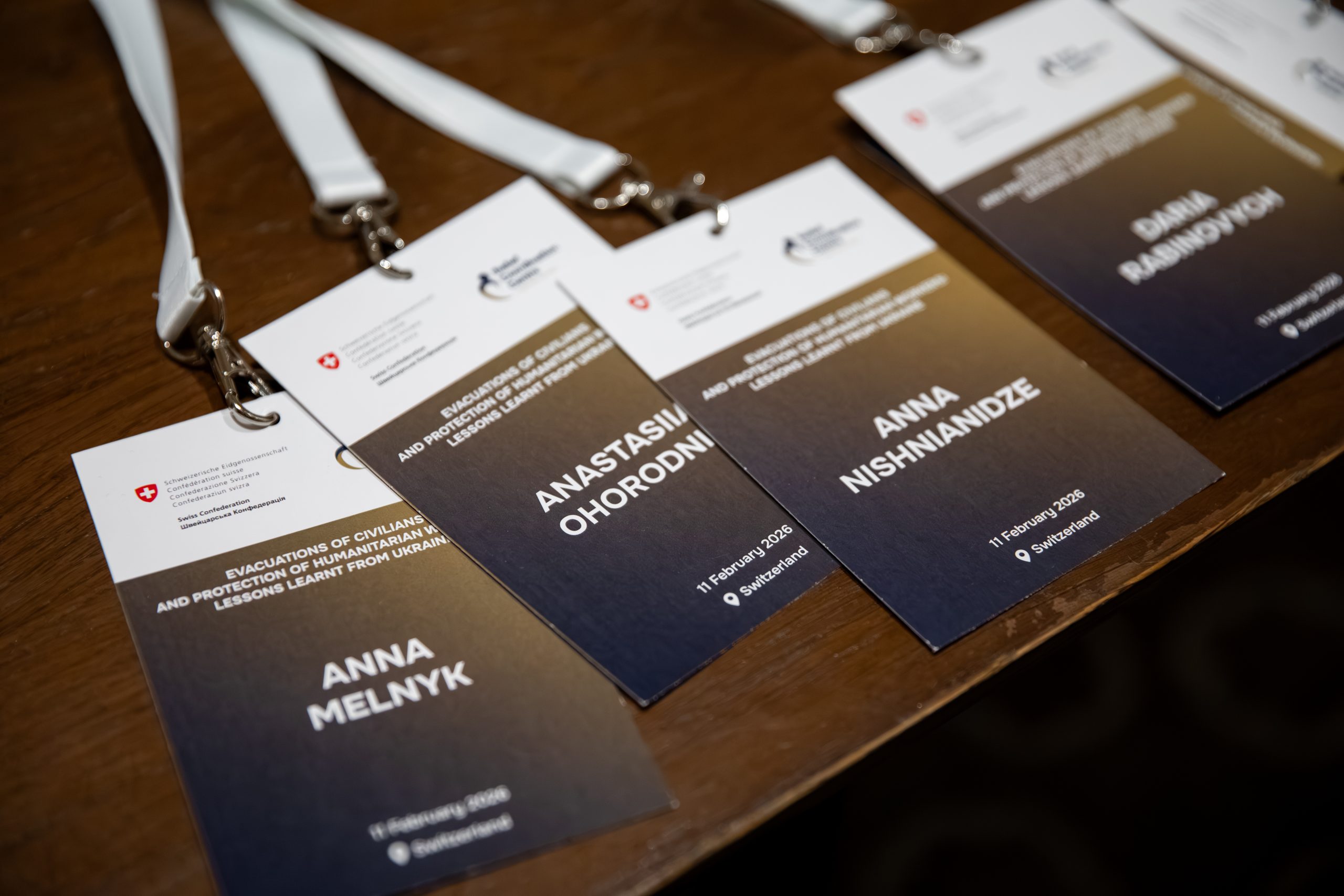 Іменні бейджі з текстом "Evacuations of civilians and protection of humanitarian workers: lessons learnt from Ukraine", датою 11 лютого 2026, країною Швейцарія на дерев’яному столі.
