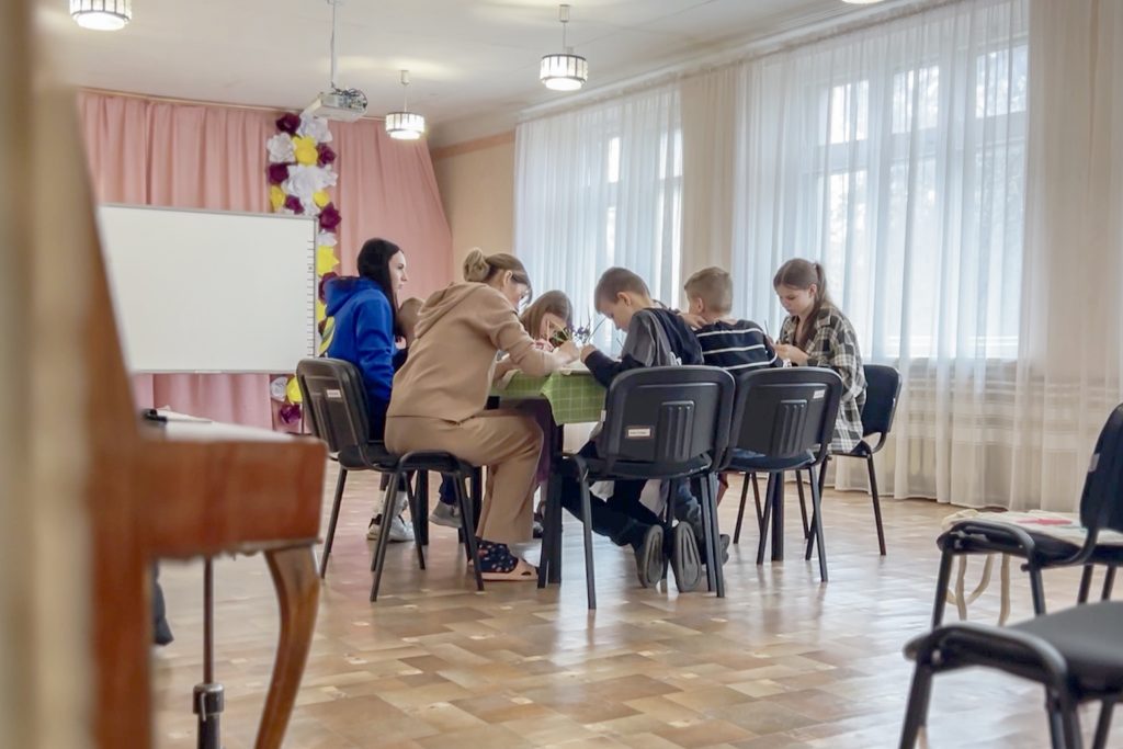 Група дітей і дорослих сидять за столом у класі з великими вікнами і розглядають або малюють щось разом.