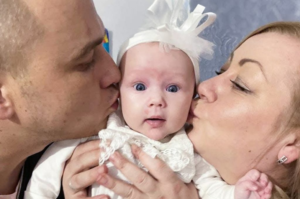 Дитина в білому одязі зі стрічкою на голові, яку цілують у щоки чоловік і жінка.