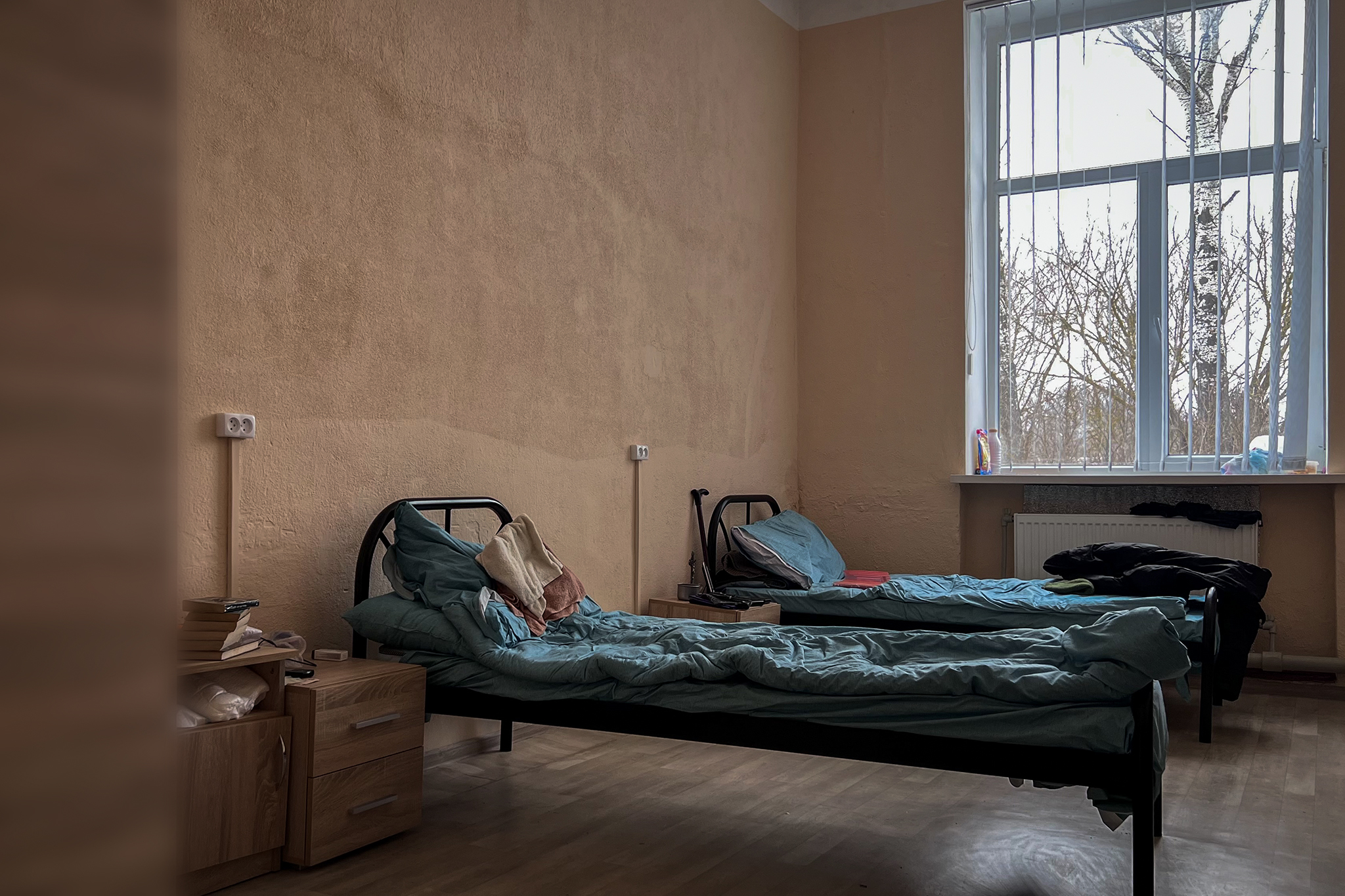 Скромна кімната з двома металевими ліжками із синім постіллю і невеликими тумбами біля стін.