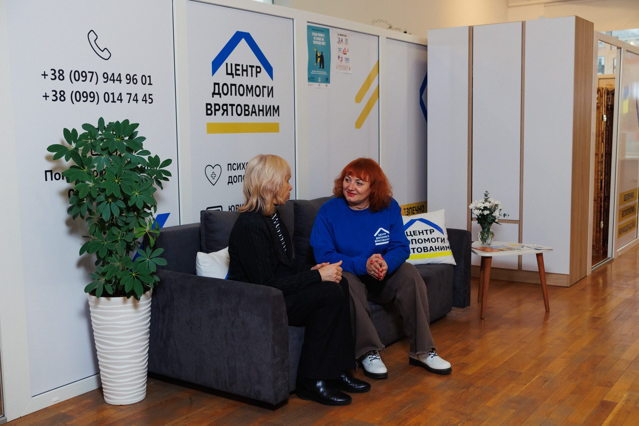 Жінки розмовляють на дивані в офісі Центру допомоги врятованим із контактами на стіні і великим зеленим квітковим горщиком поруч.
