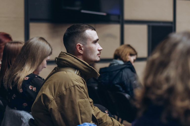 Молодий чоловік у коричневій куртці уважно дивиться вперед серед сидячих людей у приміщенні.