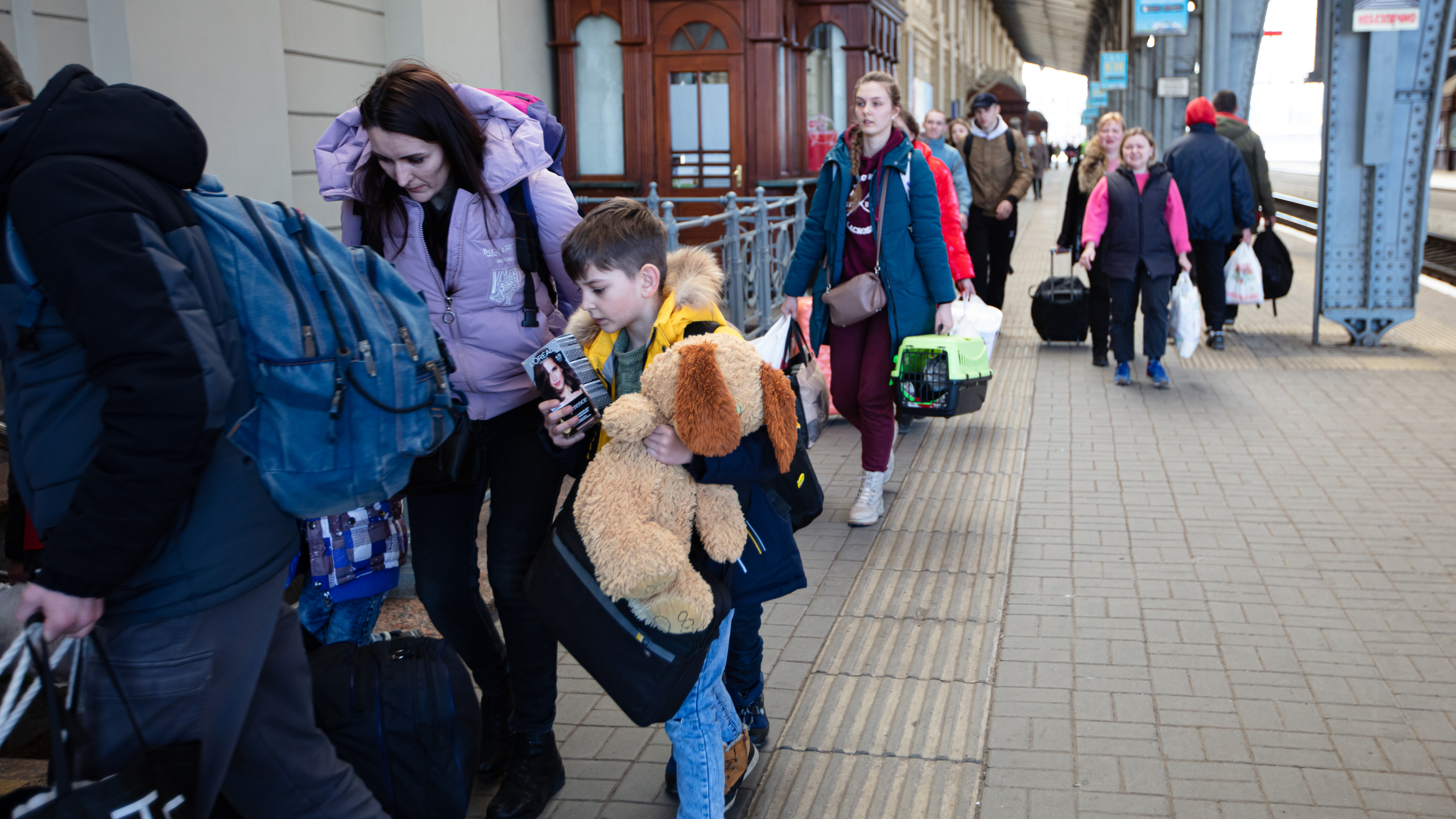 Група людей з валізами і сумками йде платформою залізничного вокзалу, хлопчик тримає великого плюшевого пса.