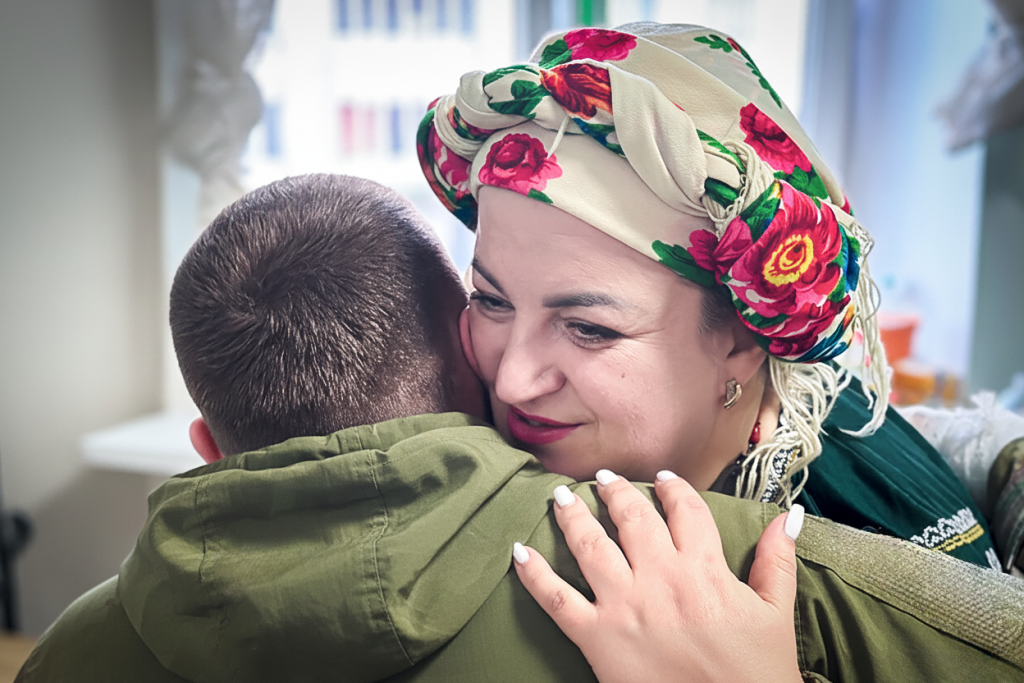 Жінка в квітчастій хустці тепло обіймає чоловіка в зеленій куртці в світлій кімнаті біля вікна.