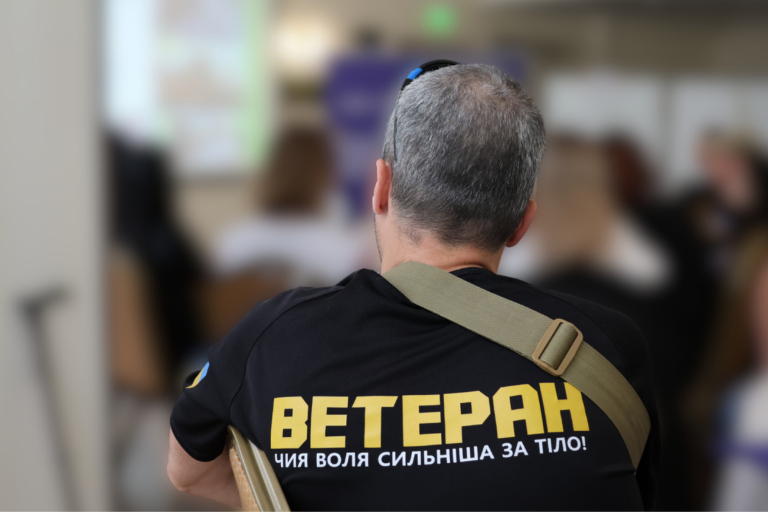Чоловік із сивим волоссям у чорній футболці з написом "ВЕТЕРАН" із жовтими літерами і фразою "ЧИЯ ВОЛЯ СИЛЬНІША ЗА ТІЛО!" на спині, у кімнаті з розмитим фоном.
