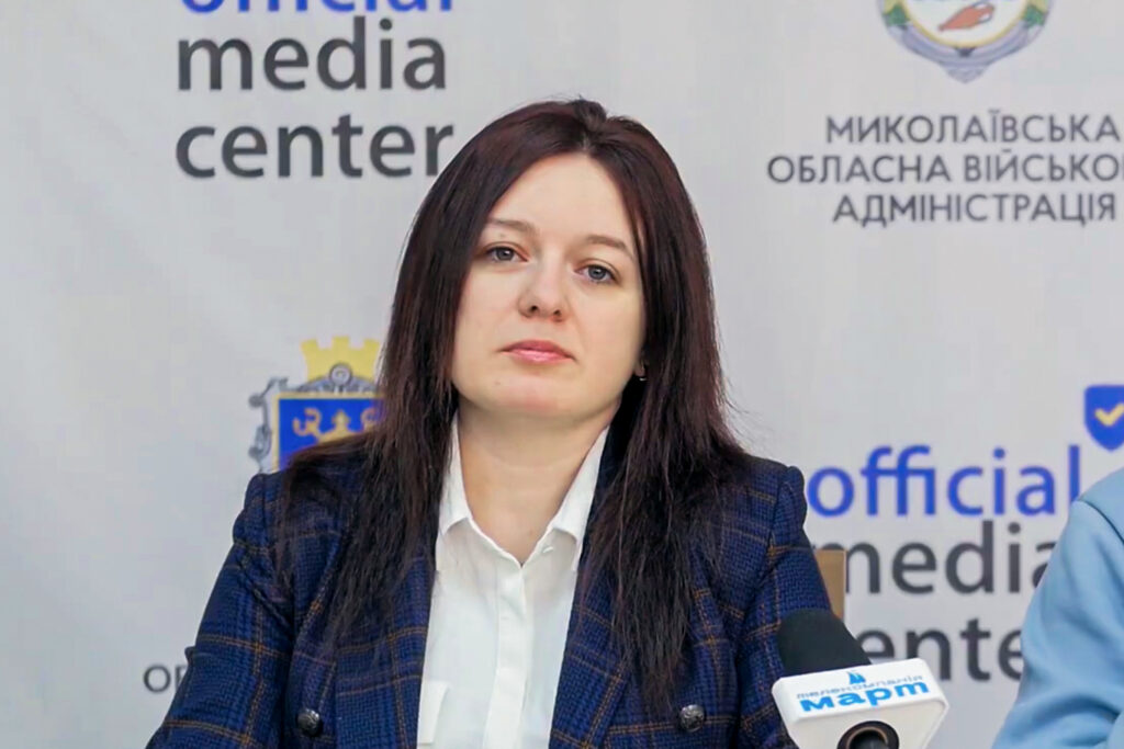 Жінка з темним довгим волоссям у синьому піджаку та білій сорочці сидить на фоні банера з написами українською, тримає мікрофон.