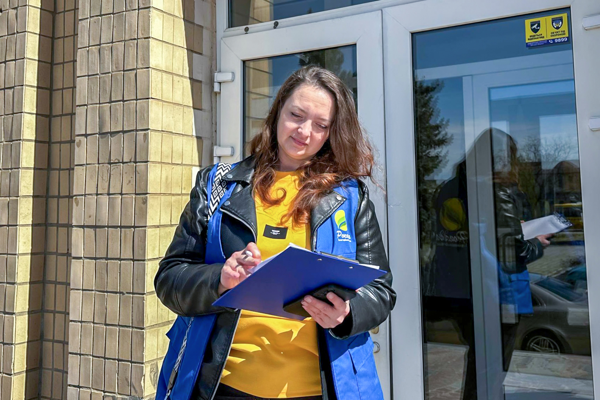 Жінка в жовтому светрі та чорній куртці і синьому жилеті  з написом "Рокада Благодійний фонд" стоїть біля вхідних дверей, тримає планшет і ручку.